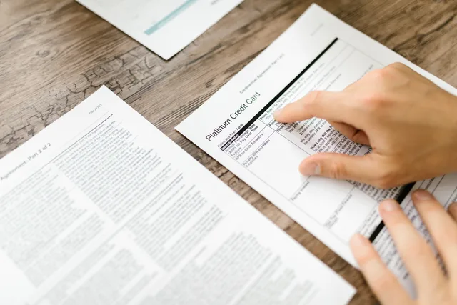 A hand examining a credit card agreement on a wooden desk, highlighting financial review. Ecommerce platforms competitor research: evidence checklist