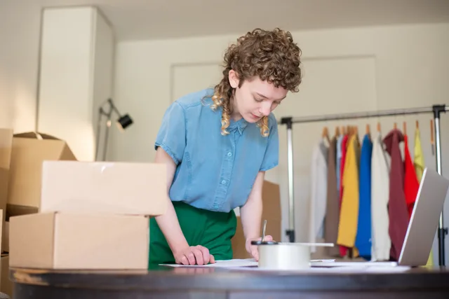 Woman packing online shop orders at home. Features boxes, clothing rack, and a laptop. Ecommerce platforms measurement: matching order, payment and settlement records across platforms and providers