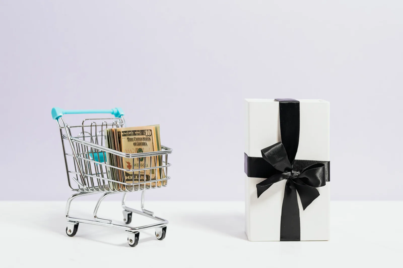 Shopping cart filled with dollar bills next to a black ribbon gift box, symbolizing commerce and presents.