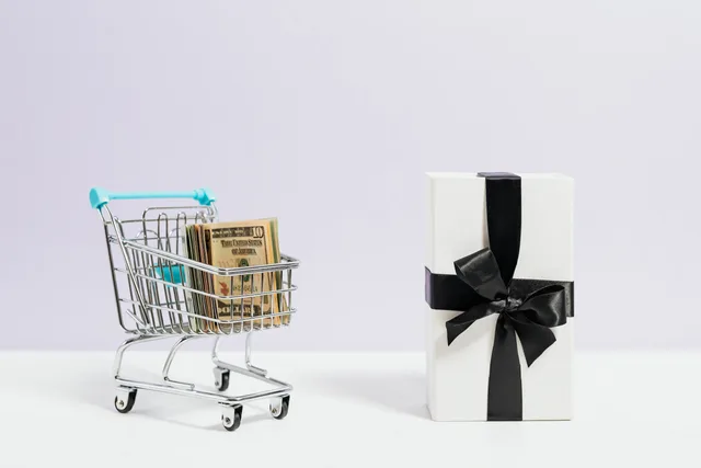 Shopping cart filled with dollar bills next to a black ribbon gift box, symbolizing commerce and presents. Ecommerce refunds: what happens to the original card fee